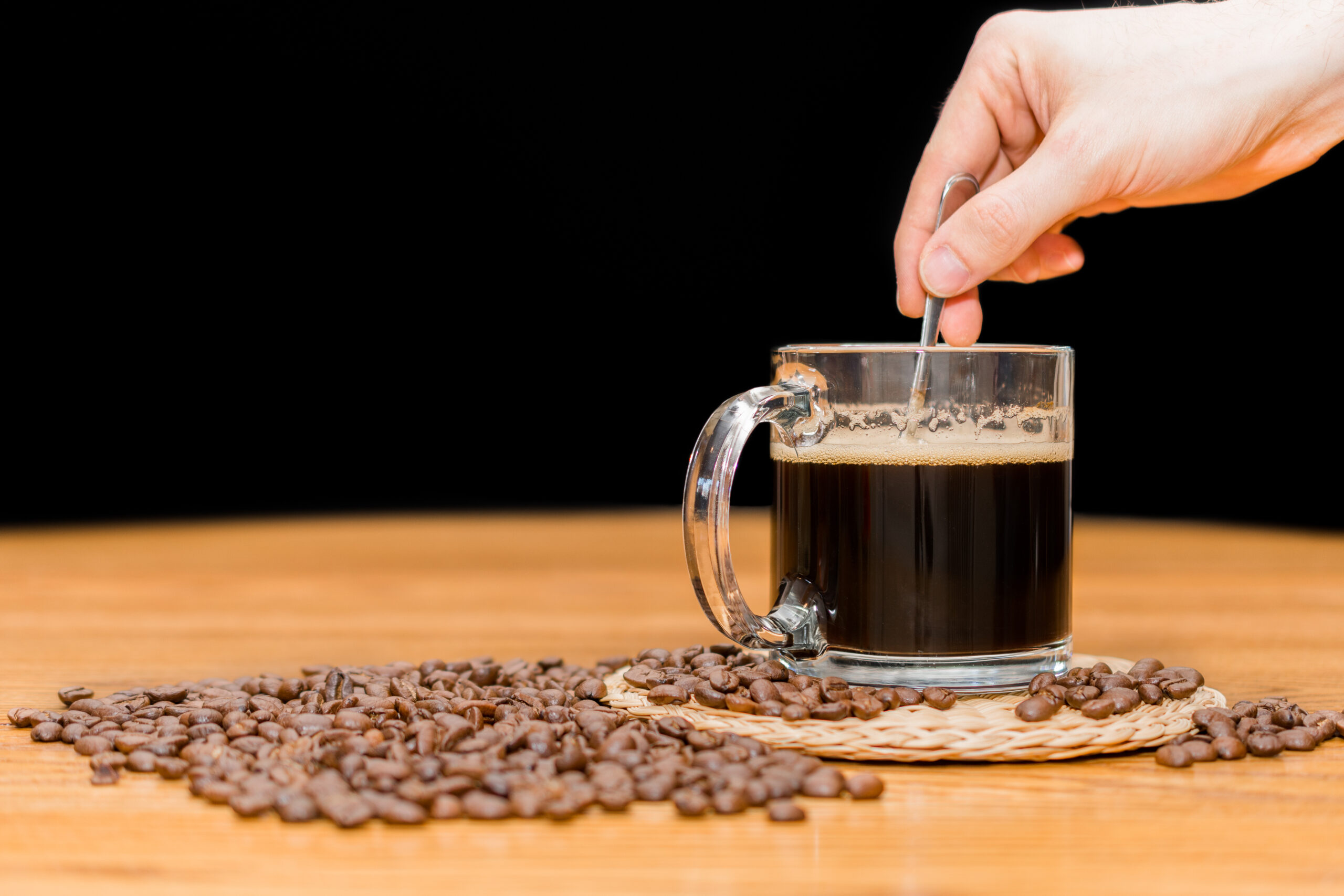 Man stirring a cup of coffee with a spoon. Coffee mug on wooden table and pile of coffee beans. isolated on black background with copy space for text